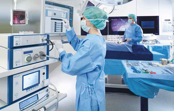 Im Bild sind zwei Mitarbeiter in OP-Kleidung zu sehen. Eine Person bedient ein medizinisches Gerät, während die andere im Hintergrund vor einem Tisch mit chirurgischen Instrumenten steht. Der Raum ist hell und modern gestaltet, mit Monitoren und weiteren medizinischen Geräten.