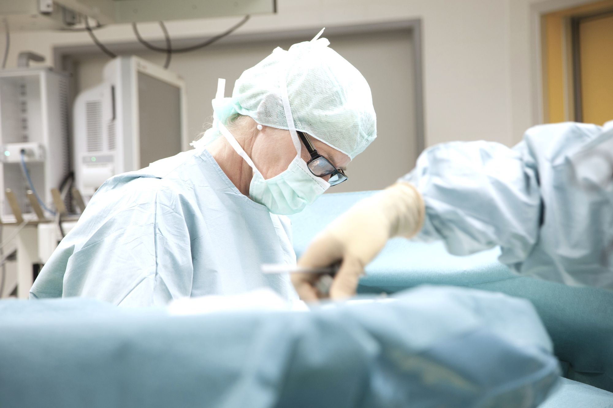 A surgical team in scrubs and masks is focused on a procedure in an operating room. The primary subject is a surgeon, wearing glasses and a surgical cap, demonstrating concentration while performing the task at hand. Bright lighting illuminates the sterile environment.