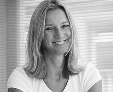 Eine lächelnde Frau mit langen, blonden Haaren trägt ein weißes T-Shirt. Sie sitzt vor einem hellen Hintergrund mit horizontalen Jalousien. Die Atmosphäre wirkt freundlich und einladend.