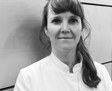 Eine Frau mit mittellangen, lockigen Haaren trägt eine weiße Arzt- oder Arbeitskleidung. Sie steht vor einer hellen Wand mit Holzpaneelen und lächelt freundlich.