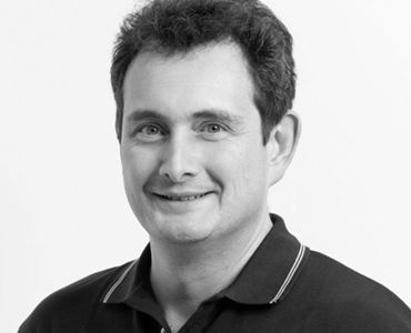 Ein Mann mit kurzem, lockigem Haar und einem freundlichen Lächeln trägt ein dunkles Polo-Shirt. Der Hintergrund ist hell und neutral. Der Fokus liegt auf seinem Gesicht und Ausdruck.