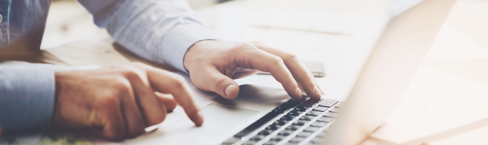 A person's hands are typing on a laptop keyboard. One hand is positioned over the keys while the other hovers nearby, focusing on the screen. The setting appears to be a workspace with a light background.