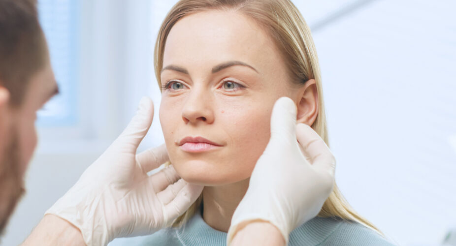 Beratung in der Plastischen und Ästhetischen Chirurgie Ein Arzt mit Handschuhen untersucht das Gesicht einer jungen Frau. Sie sitzt entspannt und schaut ihn an, während er sanft ihre Wangen hält. Der Hintergrund ist hell und klinisch, was auf eine medizinische Umgebung hinweist.