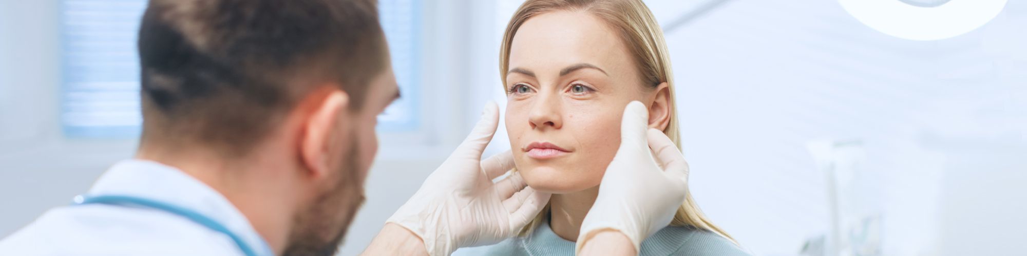 A healthcare professional in gloves examines a woman's face closely in a clinical setting. The woman appears calm as the professional assesses her features, highlighting a consultation focused on skincare or cosmetic treatment. Sunlight filters in through a window, creating a bright atmosphere.