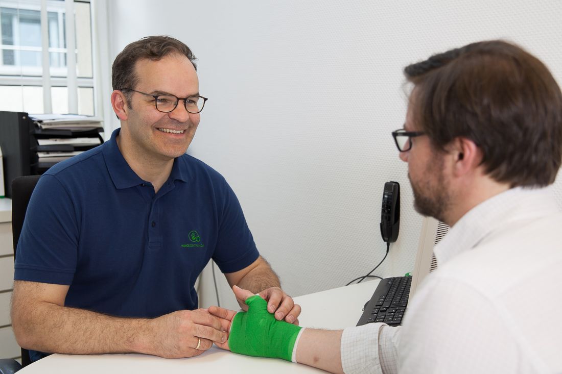 Ein Arzt sitzt an einem Schreibtisch und lächelt, während er die Hand eines Patienten mit grünem Verband betrachtet. Der Patient sieht besorgt aus. Die Umgebung wirkt hell und freundlich, was eine entspannte Atmosphäre vermittelt.