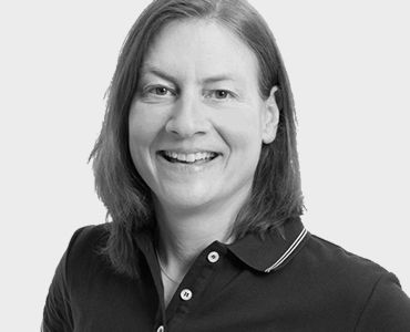 Ein Porträt einer lächelnden Frau mit mittellangen, braunen Haaren, die ein schwarzes Poloshirt trägt. Der Hintergrund ist schlicht und einfarbig. Die Frau wirkt freundlich und offen.