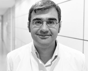 Ein Mann mit Brille und einem weißen Kittel lächelt freundlich in die Kamera. Er steht in einem hellen Korridor mit grauen Wänden.