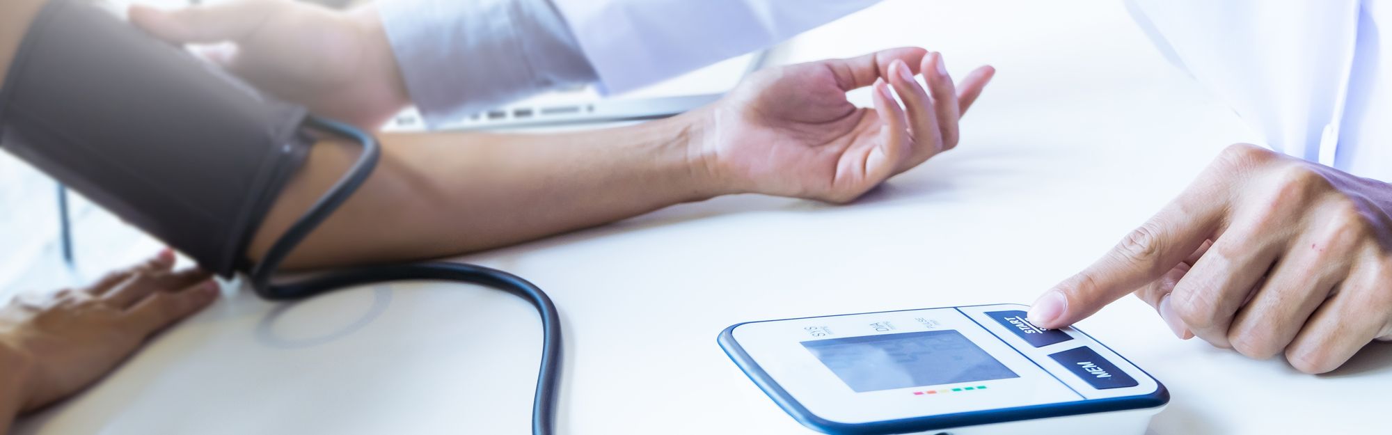 A medical professional is measuring blood pressure using an automatic device. The patient's arm is resting on a table, and the device displays readings. The healthcare provider is preparing to take the measurement by activating the device with their finger.