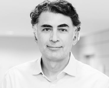 Ein Mann mit dunklen, lockigen Haaren und einem freundlichen Gesichtsausdruck steht vor einem unscharfen Hintergrund. Er trägt ein helles Hemd und hat eine aufrechte Haltung, die Selbstbewusstsein vermittelt.