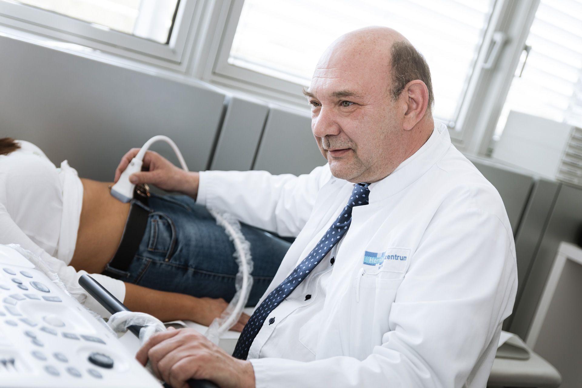 A medical professional conducts an ultrasound examination on a patient lying on an examination table. The doctor appears focused, holding the ultrasound device against the patient's side, while operating a machine nearby. The setting is a clinical environment with natural light.