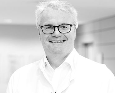 Ein lächelnder Arzt trägt eine Brille und einen weißen Kittel. Er steht in einem hellen, modernen Krankenhausumfeld. Der Arzt vermittelt Freundlichkeit und Kompetenz.