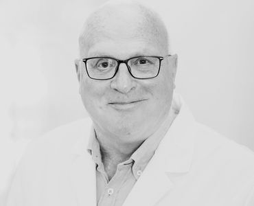 Ein lächelnder Arzt trägt eine weiße Laborjacke und Brille. Er hat eine Glatze und präsentiert einen freundlichen Ausdruck. Der Hintergrund ist unscharf und hell, was den Fokus auf das Gesicht lenkt.