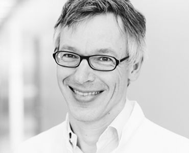 Ein lächelnder Mann mit Brille, bekleidet mit einem weißen Arztkittel. Der Hintergrund ist hell und unscharf, was den Fokus auf sein Gesicht lenkt. Der Mann vermittelt einen freundlichen und professionellen Eindruck.