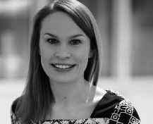 Porträt einer lächelnden Frau mit langen, glatten Haaren. Sie trägt ein gemustertes Kleid und schaut direkt in die Kamera. Der Hintergrund ist unscharf und hell.