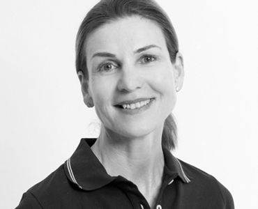 Porträt einer lächelnden Frau mit leicht zurückgehaltener Haarfrisur, die ein dunkles Poloshirt trägt. Der Hintergrund ist neutral und hell. Die Person strahlt eine freundliche und einladende Ausstrahlung aus.