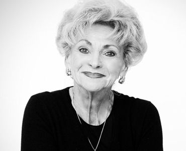 A smiling elderly woman with short, wavy hair is wearing a black top and earrings. She exudes warmth and friendliness, having a gentle expression on her face. The background is light, emphasizing her joyful demeanor.
