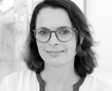 Porträt einer Frau mit Brille und einem freundlichen Lächeln. Sie hat dunkle, lockige Haare und trägt ein helles Oberteil mit einem farbigen Kragen. Der Hintergrund ist verschwommen, sodass der Fokus auf ihrem Gesicht liegt.