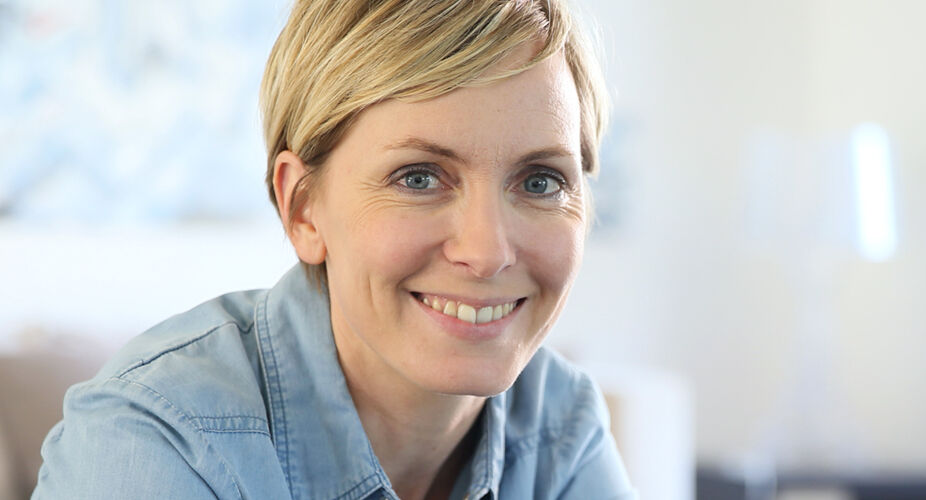 A woman with short blonde hair smiles warmly at the camera. She is wearing a denim shirt and appears to be sitting in a well-lit, comfortable indoor space. The atmosphere seems relaxed and inviting.