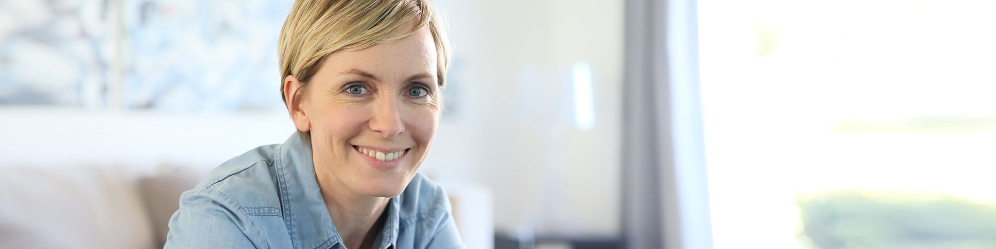 A woman with short blonde hair smiles warmly at the camera. She is wearing a denim shirt and appears to be sitting in a well-lit, comfortable indoor space. The atmosphere seems relaxed and inviting.