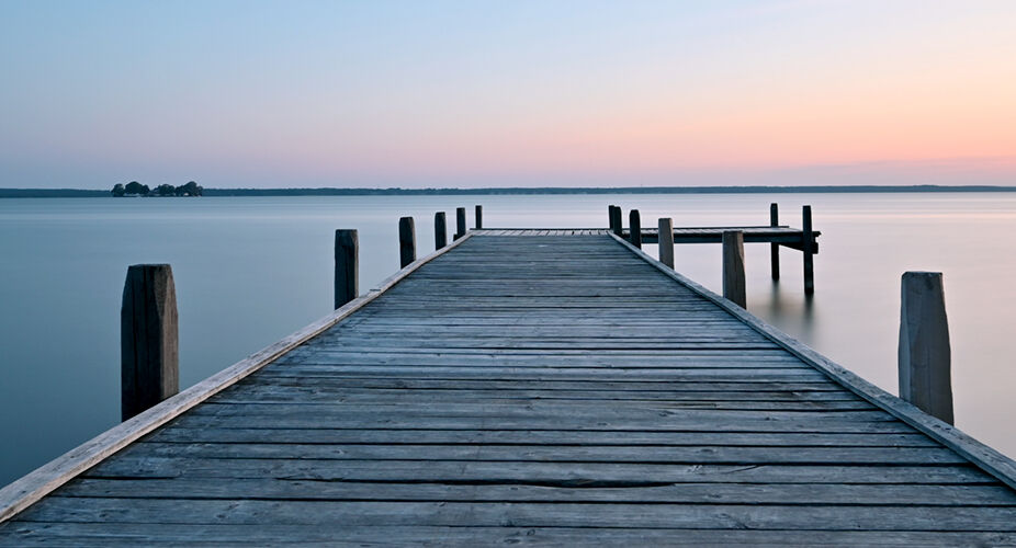Psychosomatik – Wege zu mehr Balance und Wohlbefinden Ein hölzerner Steg erstreckt sich über das ruhige Wasser, im Hintergrund ist der Horizont mit sanften Farben des Sonnenuntergangs zu sehen. Die Atmosphäre vermittelt Ruhe und Frieden.
