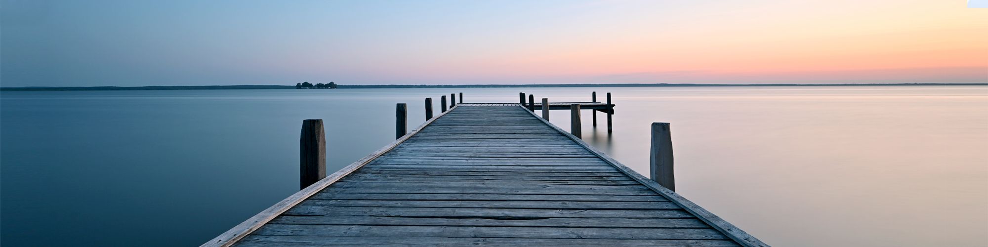 A wooden pier extends into calm water under a serene sky, transitioning from blue to soft hues of orange and pink at sunset. The scene evokes tranquility and connection with nature.