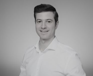 Ein lächelnder Mann mit kurzen, dunklen Haaren trägt ein weißes Hemd. Er steht in leicht seitlicher Position und blickt in die Kamera. Der Hintergrund ist einfarbig und sorgt für einen neutralen Rahmen.