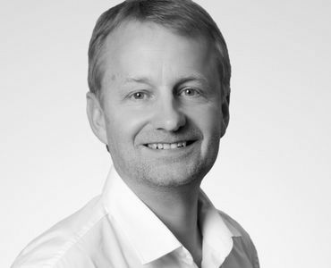 Porträt eines Mannes mit kurzem, hellblondem Haar, der in die Kamera lächelt. Er trägt ein weißes Hemd und hat einen freundlichen Ausdruck. Der Hintergrund ist neutral und hell gestaltet.