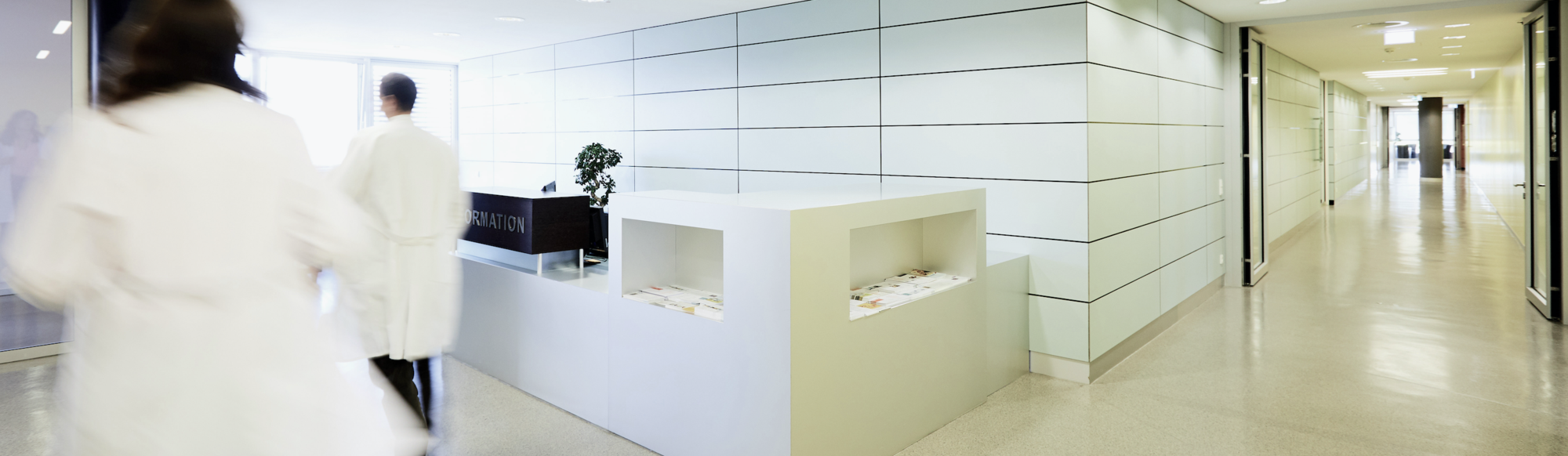 Ein moderner Krankenhausflur mit hellen Wänden und einem Empfangstresen. Zwei Personen in weißen Kitteln bewegen sich im Flur, während eine Pflanze auf dem Empfangstresen steht. Der Bereich wirkt sauber und einladend.
