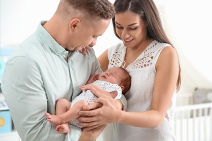 A young couple lovingly cradles their newborn baby together. The father, wearing a light green shirt, gazes at the baby, while the mother, in a white top, smiles softly. The background suggests a cozy nursery setting.