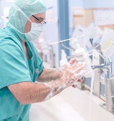 A healthcare professional in scrubs and a surgical mask washes their hands thoroughly at a sink, applying soap. The focus is on proper hand hygiene in a medical environment, emphasizing cleanliness and safety.