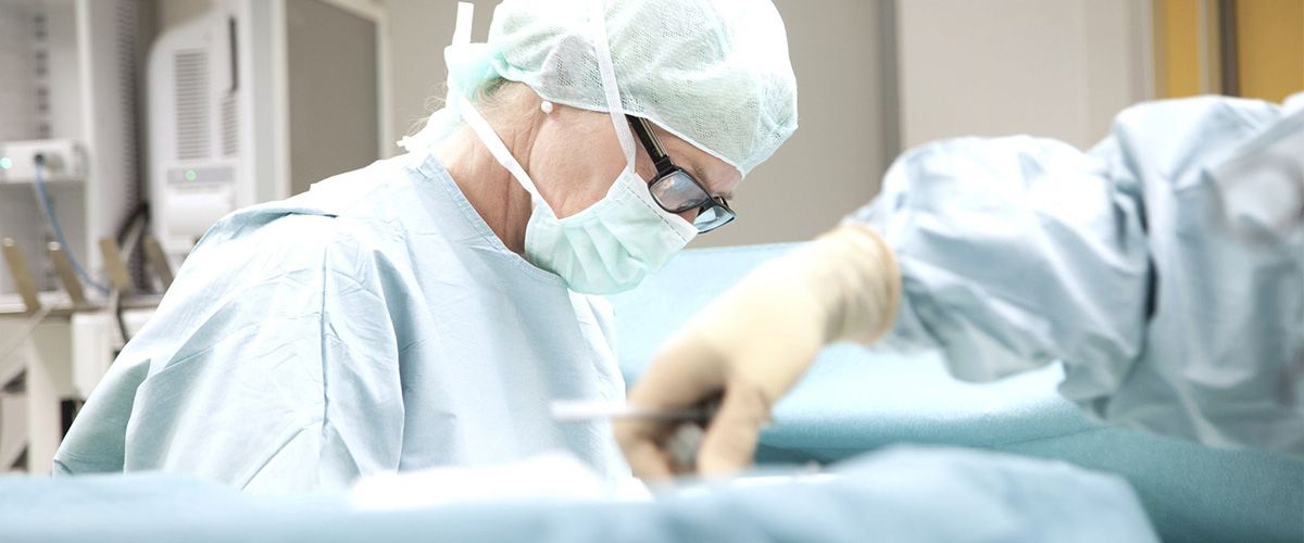 Ein Chirurg in OP-Kleidung arbeitet konzentriert an einem Patienten. Er trägt eine Schutzmaske und eine Haube, während er mit Instrumenten hantiert. Die Umgebung wirkt steril und professionell, was auf ein chirurgisches Setting hinweist.
