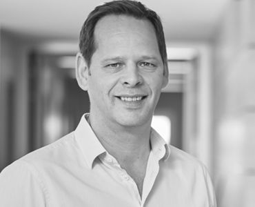 Ein lächelnder Mann mit schulterlangen, dunklen Haaren und einem hellen Hemd steht in einem Innenraum. Der Hintergrund ist verschwommen und zeigt neutrale Farben.