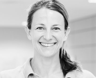 Eine lächelnde Frau mit langen Haaren, die zu einem Pferdeschwanz gebunden sind. Sie trägt ein helles Hemd und steht in einem hellen, modernen Raum. Ihr freundlicher Ausdruck vermittelt eine positive Stimmung.