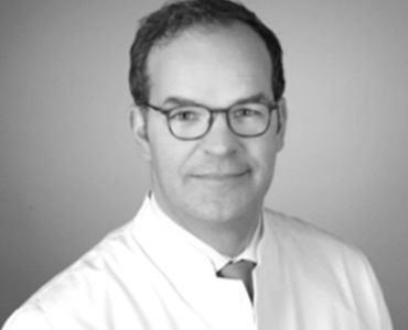 Porträt eines Arztes in einem weißen Kittel. Er trägt eine Brille und hat einen neutralen Gesichtsausdruck. Der Hintergrund ist unscharf und einfarbig.