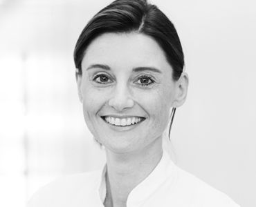 Eine lächelnde Frau trägt eine helle, medizinische Kleidung. Sie hat kurze, dunkle Haare und einen freundlichen Ausdruck. Der Hintergrund ist unscharf und hell gehalten.