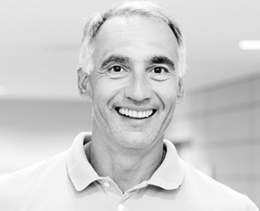 Ein lächelnder Mann mit grauen Haaren steht in einem hellen Raum. Er trägt ein helles Polo-Shirt und wirkt freundlich und einladend. Der Hintergrund ist verschwommen, wodurch der Fokus auf seinem Gesicht liegt.