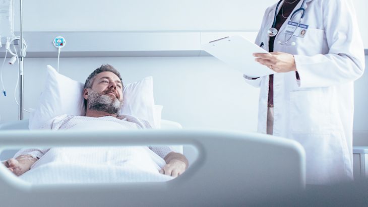 A smiling male patient lies in a hospital bed, looking at a doctor who is holding a clipboard. The setting is a bright, clean hospital room, conveying a sense of care and attention. The doctor appears attentive and engaged with the patient.