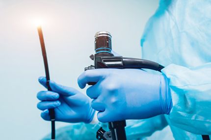 A healthcare professional wearing blue gloves holds a medical device, possibly an endoscope, in one hand and a flexible tube in the other. The setting suggests it is for a medical procedure. The background is light blue, emphasizing the sterile environment.