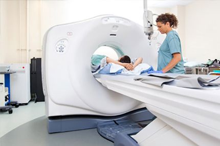 A patient lies face down on a hospital bed, preparing for a CT scan. A healthcare professional stands beside the patient, offering assistance. The CT scanner, a large, cylindrical machine, is positioned ready to perform the imaging procedure.