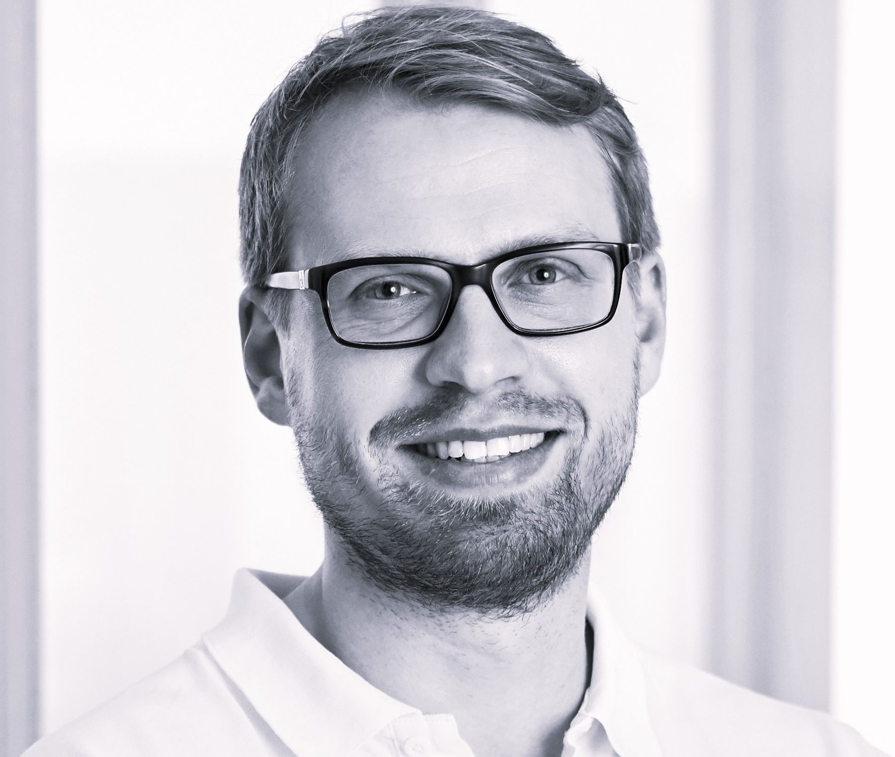 Ein lächelnder Mann mit Brille und einem Bart trägt ein weißes Poloshirt. Im Hintergrund sind unscharfe Fenster zu sehen, die auf eine helle, freundliche Umgebung hinweisen.