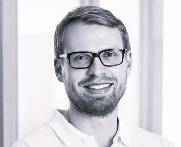 Ein lächelnder Mann mit Brille und einem Bart trägt ein weißes Poloshirt. Im Hintergrund sind unscharfe Fenster zu sehen, die auf eine helle, freundliche Umgebung hinweisen.