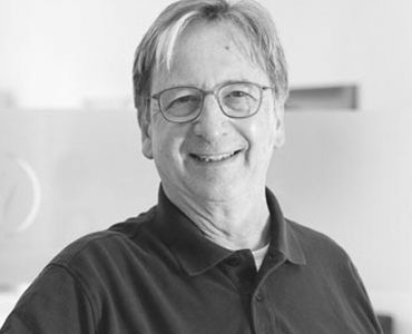 Ein lächelnder älterer Mann mit Brille trägt ein schwarzes Polo-Shirt. Er steht in einem hellen Raum und wirkt freundlich und einladend.