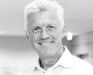 Ein älterer Mann mit grauen Haaren trägt ein weißes Hemd und lächelt freundlich in die Kamera. Der Hintergrund ist unscharf und hell.