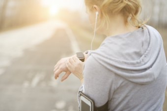 Eine Person mit Kopfhörern schaut auf ihre Smartwatch, während sie an einem leeren Weg läuft. Die Sonne scheint im Hintergrund und erzeugt eine angenehme Atmosphäre.