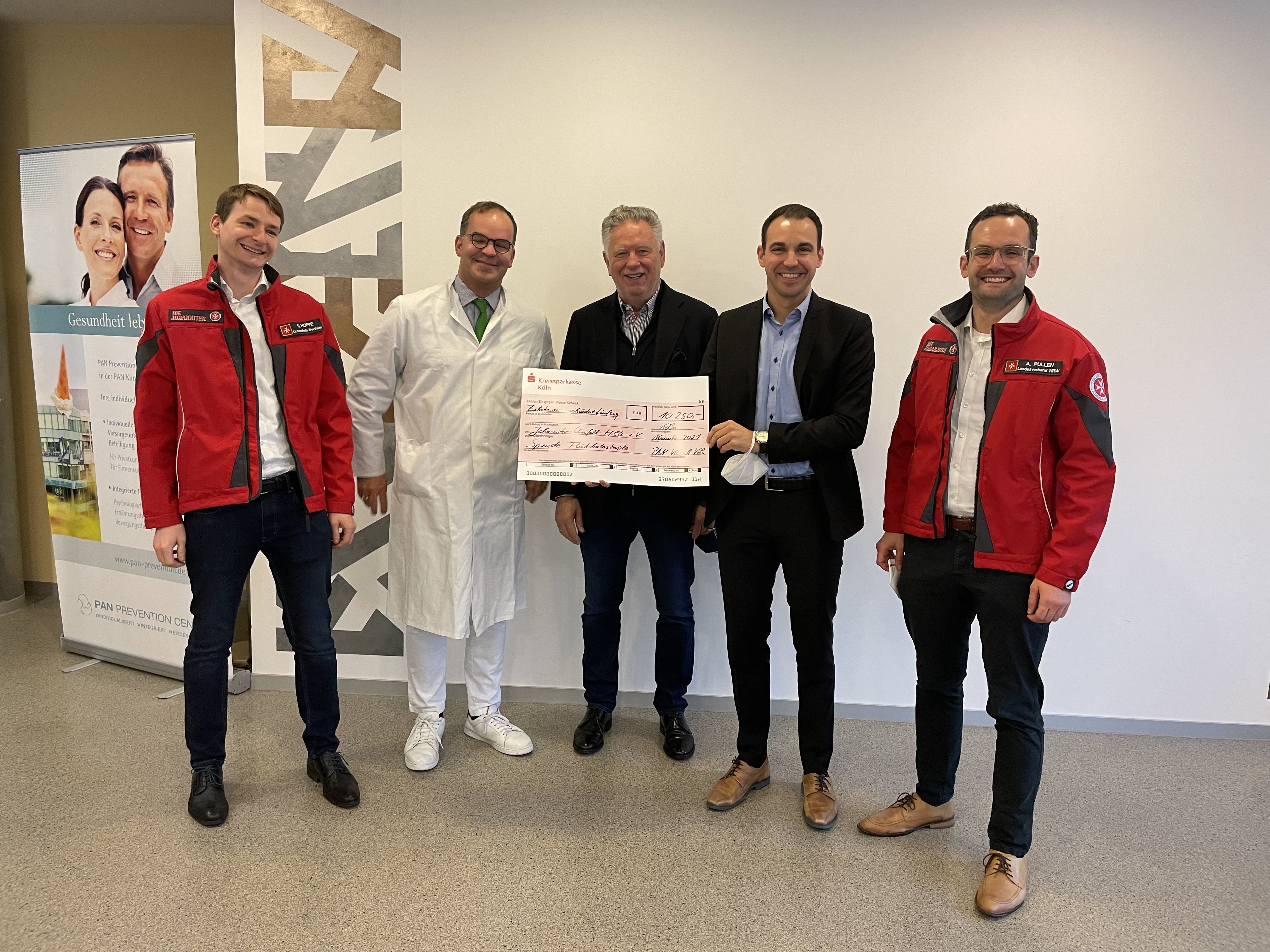 Fünf Personen stehen in einem Innenraum. Ein Mann in einem weißen Kittel übergibt einen großen Scheck. Alle Teilnehmer lächeln und tragen formelle Kleidung. Im Hintergrund ist ein Banner mit Fotografien zu sehen. Der Scheck symbolisiert eine Spende oder Unterstützung.