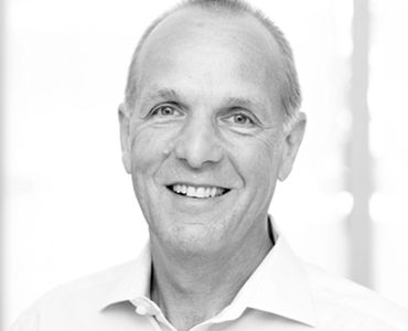 Ein lächelnder Mann mit kurzem, grauem Haar trägt ein helles Hemd. Sein Gesicht zeigt Freundlichkeit und Offenheit. Der Hintergrund ist hell und unscharf, wodurch die Aufmerksamkeit auf seinen Ausdruck gelenkt wird.