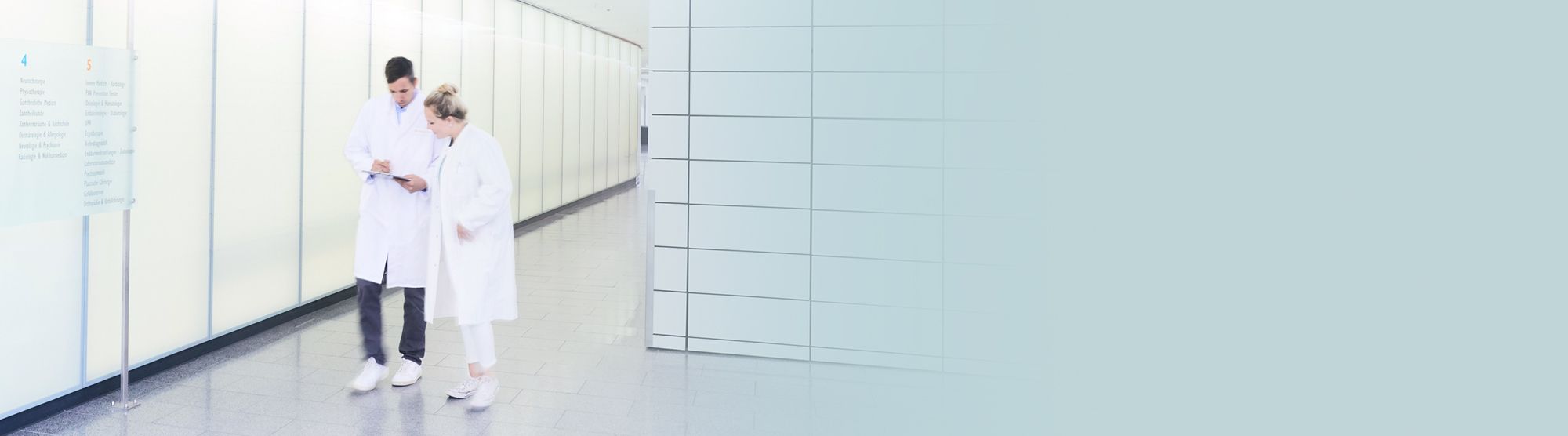 Two healthcare professionals in white lab coats are discussing information while walking down a modern hallway with bright, white walls. One is holding a digital device, likely a tablet or phone, as they collaborate.