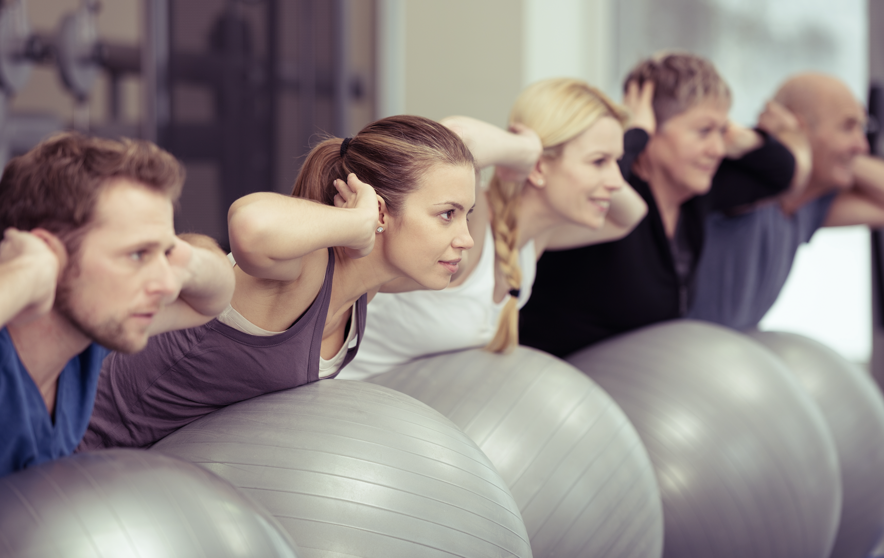 Eine Gruppe von fünf Menschen macht Gymnastik auf großen Fitnessbällen. Sie lehnen sich nach vorne und halten den Kopf mit den Händen, während sie die Übung durchführen. Die Atmosphäre wirkt konzentriert und aktiv.
