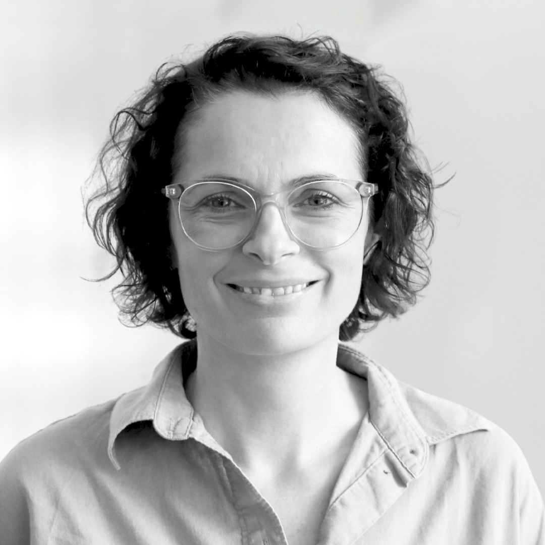 Ein Portrait einer lächelnden Frau mit kurzen, lockigen Haaren und einer gleichmäßigen Brille. Sie trägt ein helles Hemd und hat einen freundlichen Ausdruck. Der Hintergrund ist neutral und unscharf, wodurch der Fokus auf ihrem Gesicht liegt.