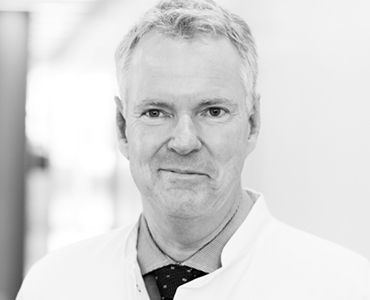 Ein Mann in einem weißen Laborkittel schaut freundlich in die Kamera. Er hat kurzes, graues Haar und trägt ein Hemd mit Krawatte. Der Hintergrund ist unscharf, was den Fokus auf sein Gesicht legt.